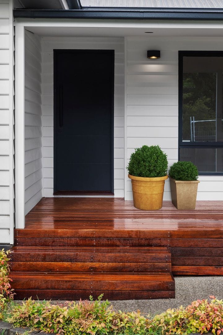 Front entrance of a house with a black door, wooden deck steps, potted plants, and white siding.