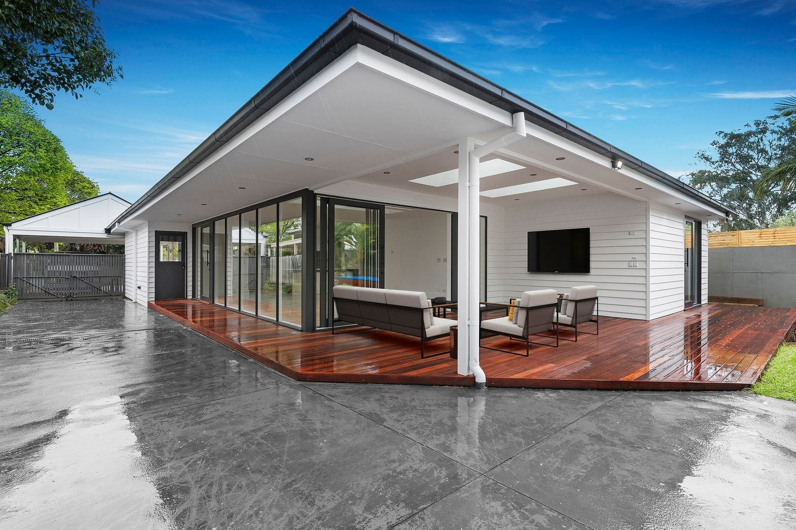 Modern single-story house with large glass doors, covered patio, wooden decking, and outdoor seating.