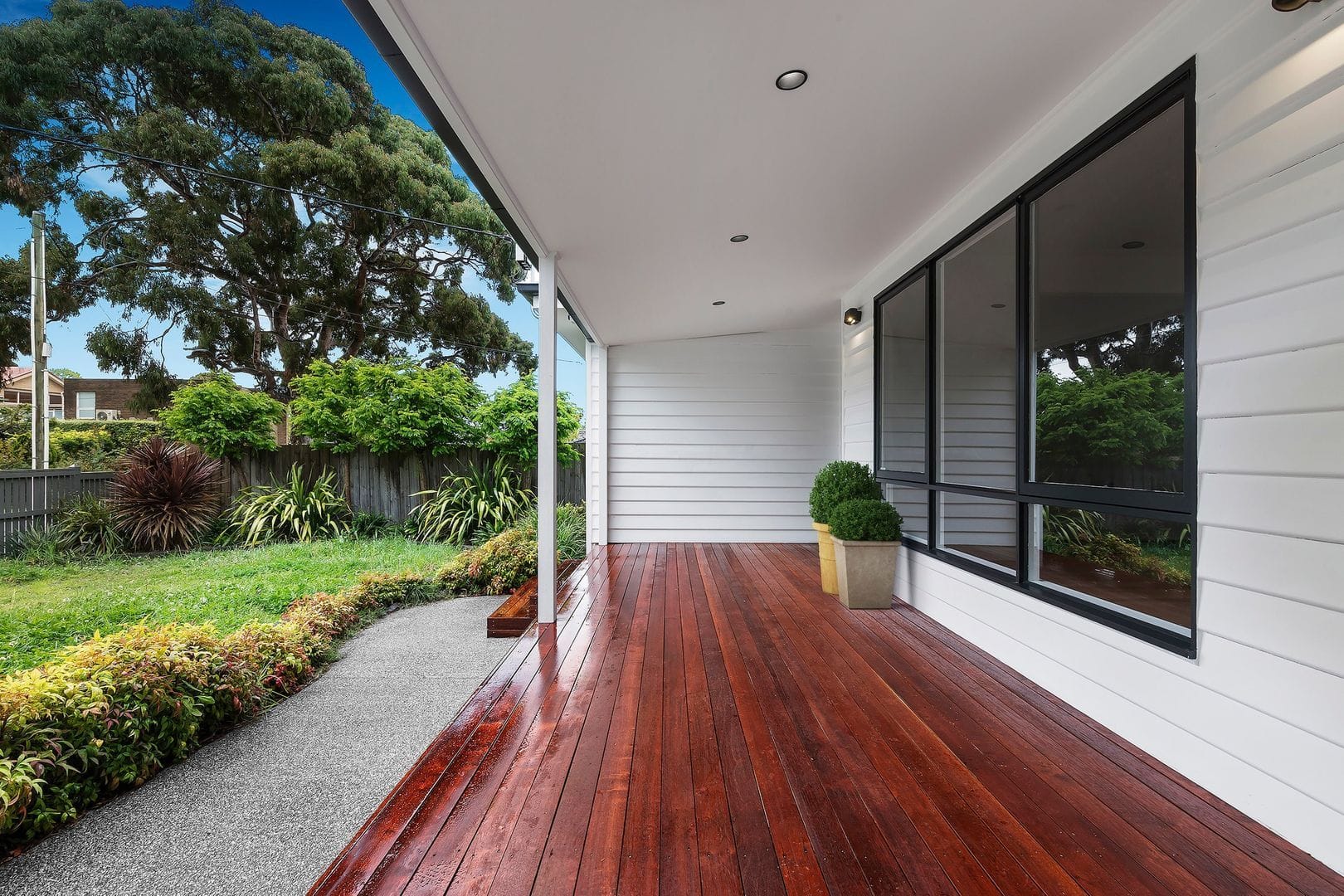 Polished wooden deck attached to a house with white siding, large windows, recessed lighting, and a lush garden.