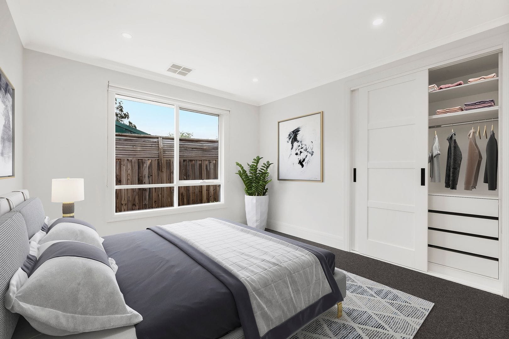 Modern bedroom featuring a bed with gray and white bedding, a bedside lamp, large window, abstract wall art, potted plant, and an open closet.