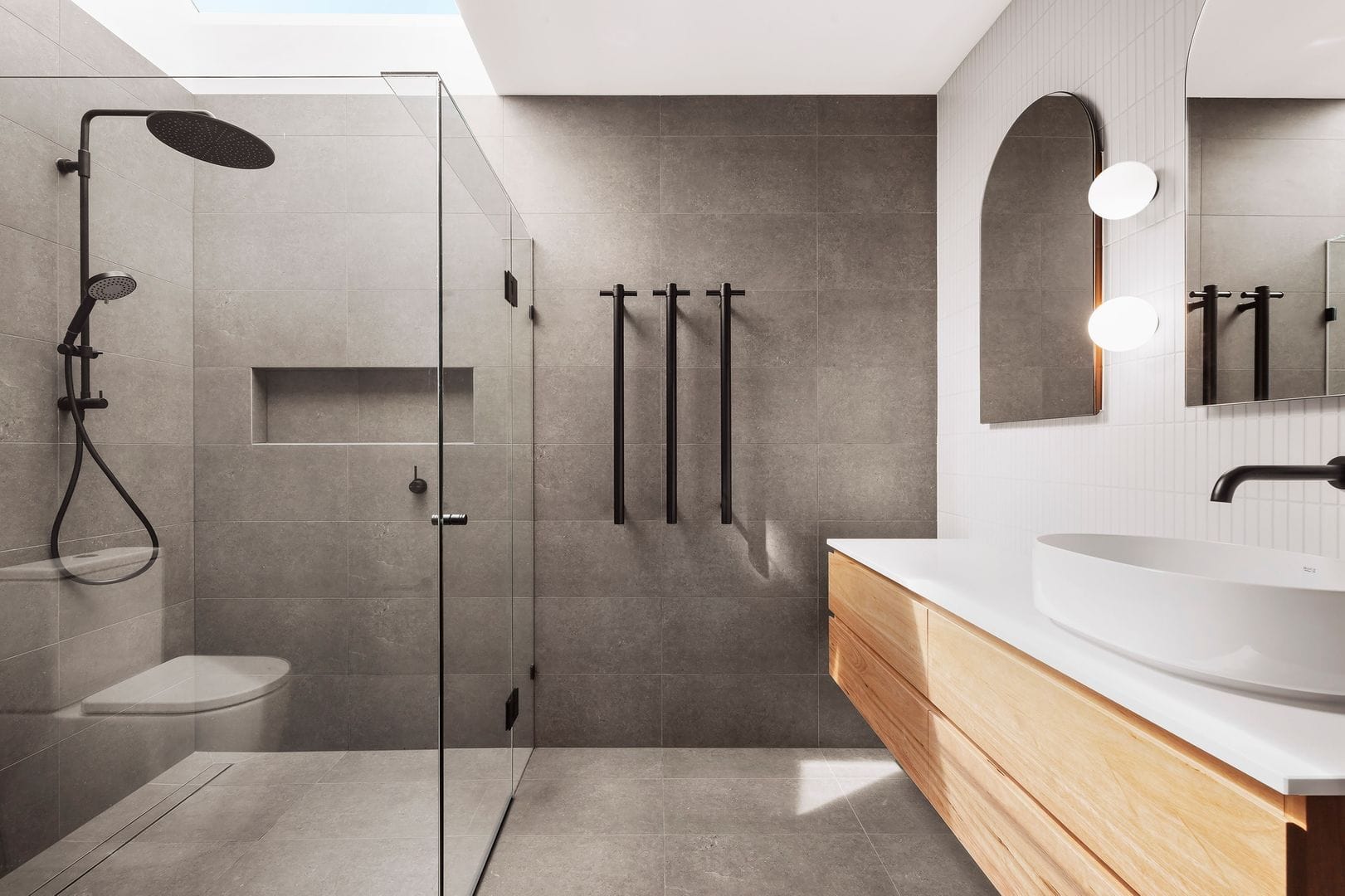Modern bathroom with glass-enclosed shower, floating wooden vanity, grey tiles, and natural lighting.