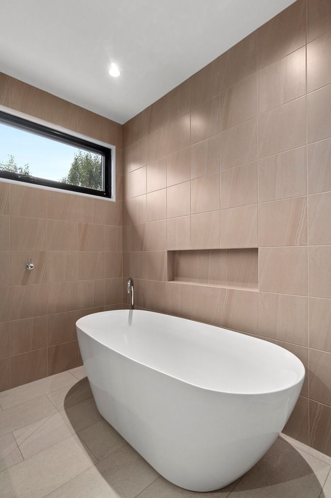 Modern bathroom with freestanding white bathtub, beige tiles, recessed shelf, and small window.