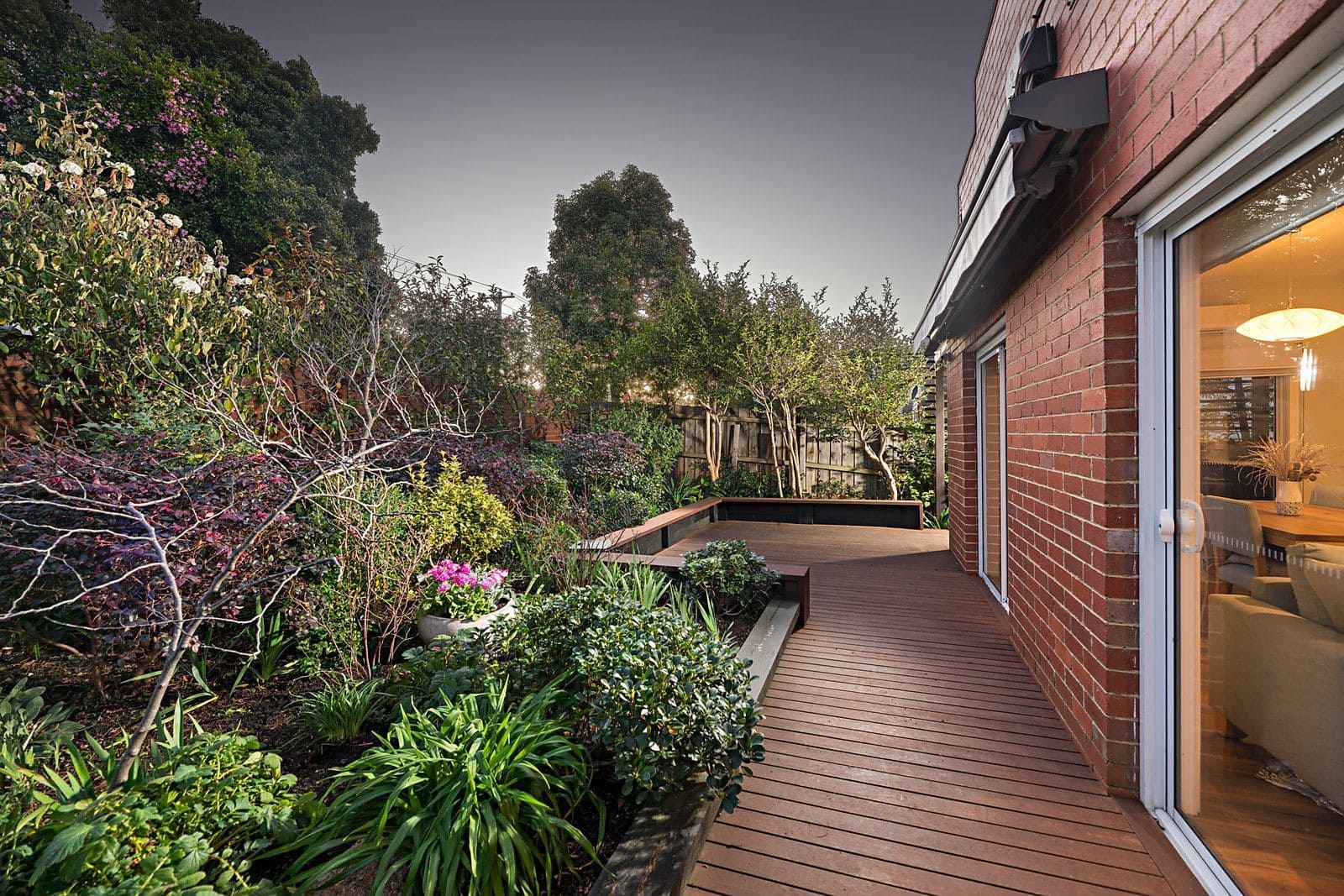 Wooden deck pathway beside red brick house with lush garden and sliding glass doors.