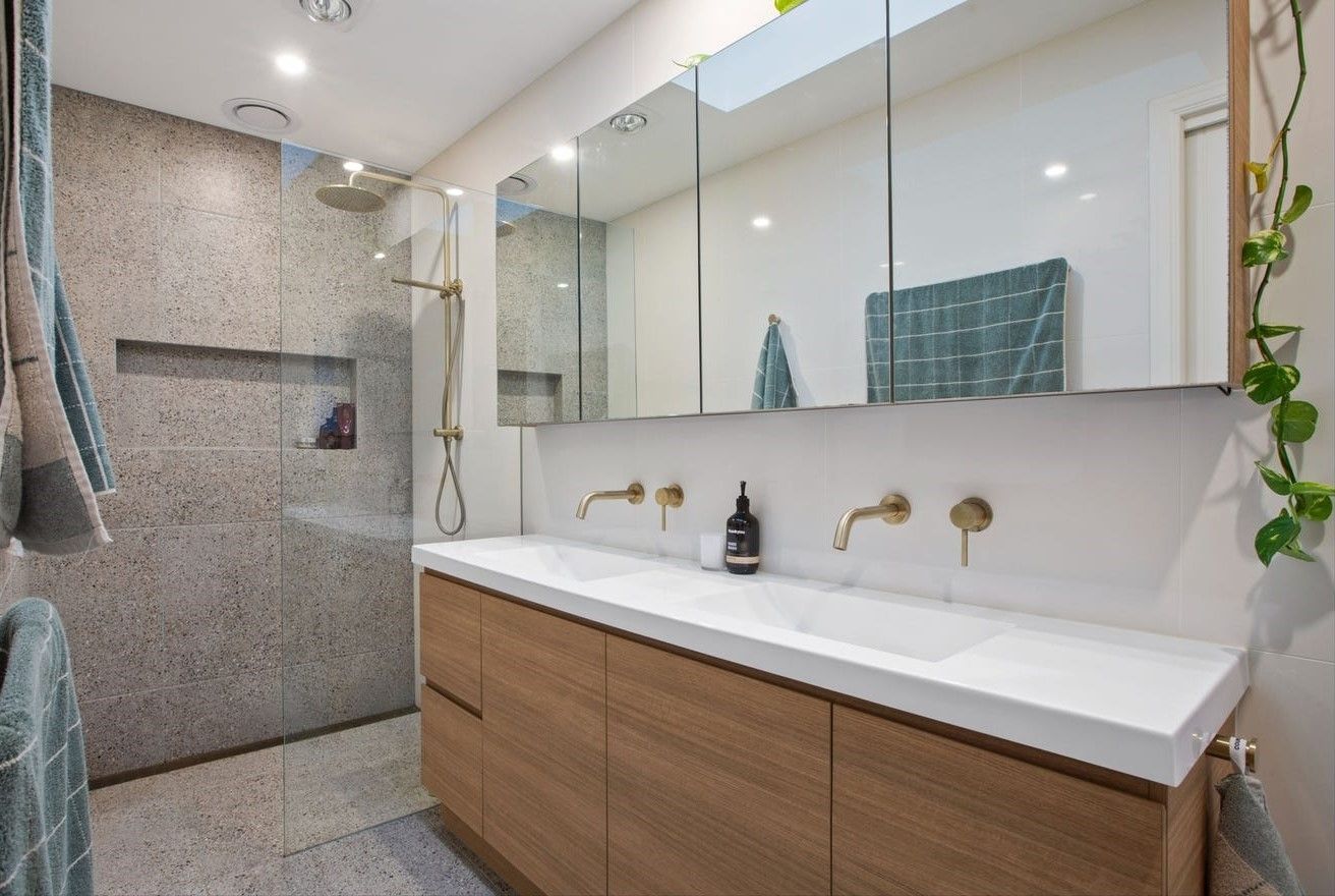 A modern bathroom with a double vanity, gold faucets, large mirrors, and a glass-enclosed shower area.