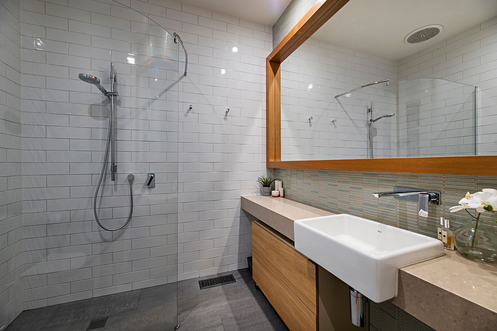 Modern bathroom with white subway tiles, glass shower partition, large mirror, beige countertop, and wooden cabinetry.